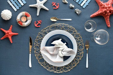 Columbus Day or Summer birthday lunch table, blue white with red. Flat lay, contemporary nautical table setting with sea decorations on classic blue linen textile. Plates, golden cutlery, sea shells