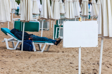Fototapeta na wymiar Information board in beach area. A warmly dressed man sleeps on a deck chair next to closed umbrellas. Selective focus, place for text. The beginning of the tourist season. Summer holiday concept.