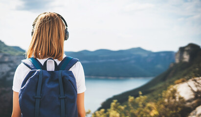 back view girl backpacker with light hair dreams listening to music in headphones relaxation on top rocky mountains, blonde hipster with backpack leisure after walking summer lake valley, copy space