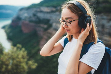 relaxed young female traveler hipster glasses meditates with eyes closed listening to favorite music on wireless headphones while standing on top of mountain and enjoying clean air