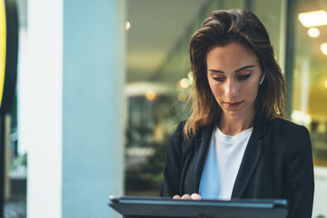 Lifestyle business woman using tablet walking in evening city, girl banker dressed in business style listening to music wireless earphones and typing text message on laptop device, flare light