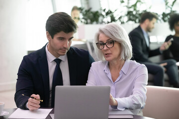 Fototapeta na wymiar Mature businesswoman use laptop to discuss information with younger colleage. Experienced business woman mentor explain strategy to young manager. Two businesspeople work with online data together