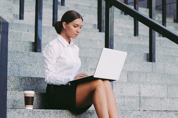 Young attractive girl student in suit sit on stairs and use laptop and drink coffee outdoors. Beautiful confident business woman typing keyword on computer on city street. Busy freelance entrepreneur