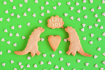 Homemade shortbread cookies decorated white glaze. Two dinosaurs with callout cloud with text ‘I love you’ on green background with heart sprinkles, top view