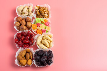 A serving of assorted candied fruit, dried cherries, almonds, raisins, walnuts and hazelnuts in paper muffin cups on a pink background. Copyspace.