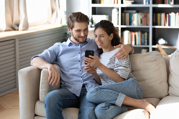 Happy young couple using smartphone together, relaxing on cozy couch in living room, smiling woman and man hugging, looking at phone screen, shopping online, spending leisure time with device
