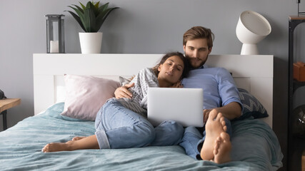 Young man and woman hugging, relaxing, lying in cozy bed in bedroom, using laptop, looking at screen, young couple watching movie, shopping online, enjoying leisure time, lazy weekend at home