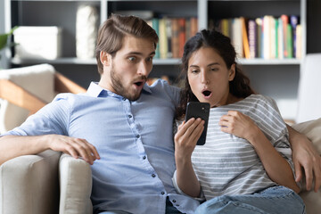 Shocked young man and woman with open mouth looking at phone screen together, reading unexpected news in message, amazed by unbelievable offer, sitting on cozy couch in living room