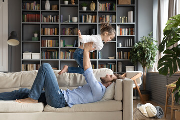 Smiling father lying on cozy couch, holding little daughter with hands outstretched pretending flying, loving young dad and cute girl playing funny game in living room, enjoying leisure time together