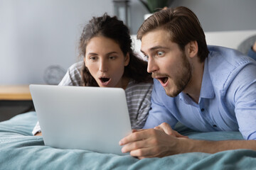 Shocked young couple looking at laptop screen, lying on bed at home, surprised woman and man with...