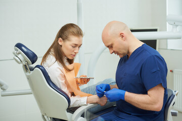 Obraz premium A bald middle-aged dentist shows a female patient tooth color samples before a whitening procedure in a dentist's office. A doctor is matching a tooth color to a patient before treatment.