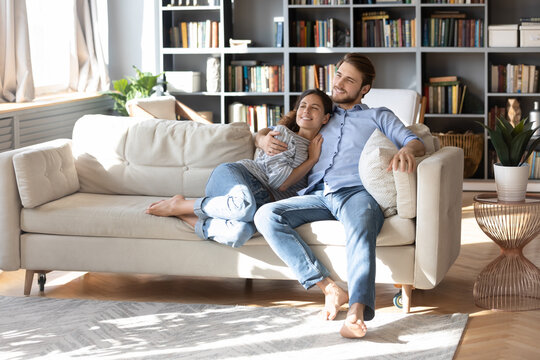 Smiling Dreamy Young Married Couple Relaxing On Cozy Couch Together, Happy Woman And Man Hugging, Looking To Aside, Family Dreaming About Good Future, Planning, Pondering New Opportunities