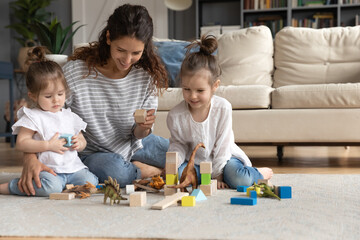 Smiling caring young mother or nanny playing with two little daughters on warm floor in modern living room at home, cute preschool girls playing with toys, colorful wooden blocks, building tower