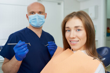 Dentist in medical face mask is holding a dental mirror and dental explorer near a smiling female...
