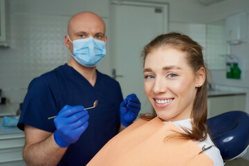 Dentist in medical face mask is holding a dental mirror and dental explorer near a laying female...