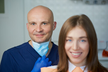 A bald smiling middle-aged dentist together with his young female patient in a dentist's office. A doctor and his patient are demonstrating an excellent result of the teeth treatment.