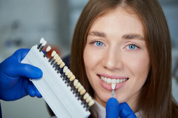 A close photo of a teeth shade sample that is matching to a smiling female patient in a dental chair. A doctor is checking teeth color samples before treatment procedure in a dentist's office.