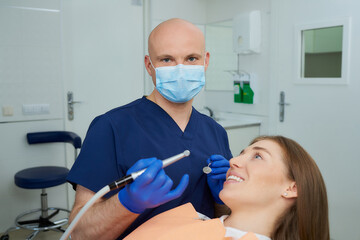 A dentist in a medical face mask holds a dental mirror and drill before a procedure near a lying young woman in the dental chair. A doctor and a female patient during treatment in a dentist's office.