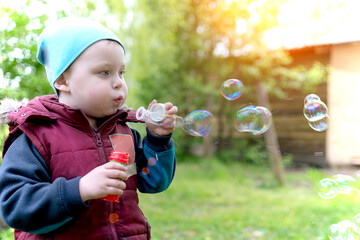 Cute little baby in spring blows soap bubbles. A boy on the street, dressed warmly. Bubbles are...