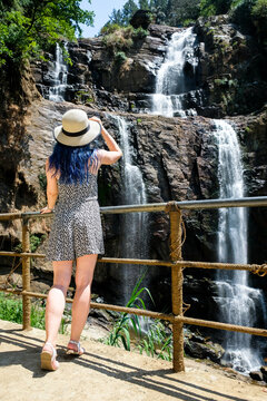 Girl with blue hair in a hat looking at a famous landmark Ramboda waterfall near Ella, Sri Lanka. View at cascade water flowing from the viewpoint on a sunny day.