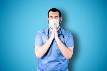 Closeup portrait young doctor with mask praying hands clasped hoping for best asking for forgiveness or miracle isolated blue wall background. Human emotion facial expression feeling