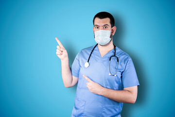 Young doctor with mask pointing and looking at copy space. Handsome young businessman isolated on blue background
