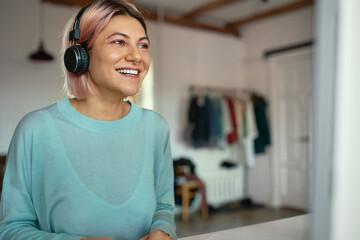 Indoor shot of beautiful happy student girl in blue sweatshirt using wireless headphones, having...