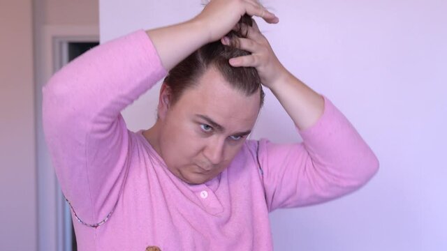 Young unshaven plump man wearing funny pink pajamas, with ridiculous long bang on one side of the head, collects the hair into one ponytail on the top. Concept of lacking of hairdresser service.