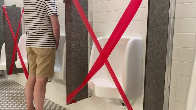 A man walks up to a urinal in a men's restroom. The urinals next to his are taped off due to social distance requirements during the COVID-19 pandemic of 2020.  	