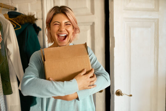 Indoor image of happy cheerful young woman holding cardboard box delivered to her apartment, expressing excitement, going to unpack parcel, having impatient overjoyed look. Food delivery and shopping