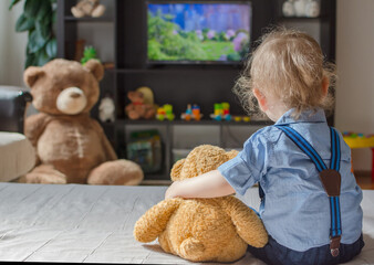 Cute baby boy and his teddy bear watching TV sitting on a couch in the living room at home