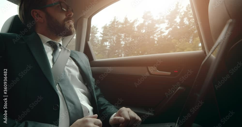 Poster thoughtful businessman in black suit and eyewear using portable computer while sitting on backseat o