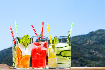 Iced summer drink. Refreshing multicolored alcol-free cocktails in glasses with straws. Watermelon,cucumber, lemon, orange, strawberry cocktail on white tray on nature mountains background.