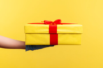 Profile side view closeup of human hand in blue surgical gloves holding yellow gift box. indoor, studio shot, isolated on yellow background.