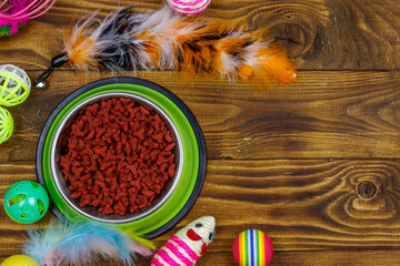 Set of toys for cat and bowl with dry pet food on wooden background. Top view, copy space. Pet care concept