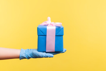 Profile side view closeup of human hand in blue surgical gloves holding blue gift box. sharing, giving or delivery concept. indoor, studio shot, isolated on yellow background.