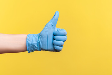 Profile side view closeup of human hand in blue surgical gloves showing like or thumbs up. indoor, studio shot, isolated on yellow background.