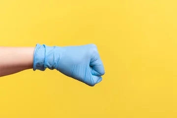 Foto op Plexiglas Profile side view closeup of human hand in blue surgical gloves showing boxing fists with hand. attack or defense against problems. indoor, studio shot, isolated on yellow background. © khosrork