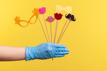 Profile side view closeup of human hand in blue surgical gloves holding and showing many different stick photo-booth in hand. indoor, studio shot, isolated on yellow background.