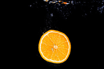 An orange falling into water on a black shiny background falls under water with bubbles.