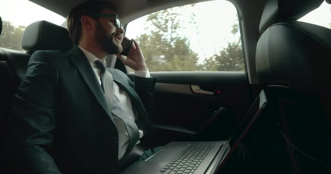 Bearded man in eyewear and business suit talking on smartphone during riding in car with opened laptop on knees. Mature businessman sitting on back seat and fastened with seatbelt.