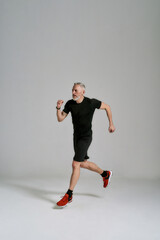 Full length shot of middle aged muscular man in black sportswear looking aside while jogging during workout in studio over grey background