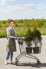 Full length portrait of smiling young woman wearing apron and looking at camera while standing by cart with tree saplings and plants outdoors, copy space
