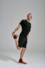 Full length shot of middle aged muscular man in black sportswear looking aside while stretching his...