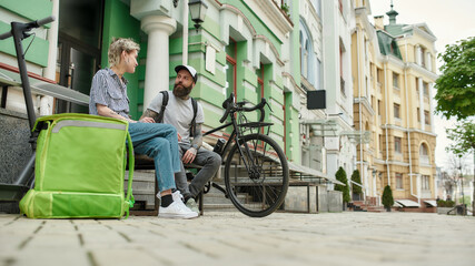 Two cheerful couriers, young man and woman sitting on the bench and talking outdoors while delivering food and products, using scooter and bike
