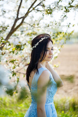 Obraz premium Beautiful girl in a light summer blue long dress adorned in her hair against a blossoming tree. Tender portrait of a young woman in white blossom