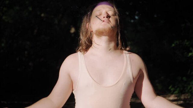 Funny Fat Man with Long Hair in a Tight T-shirt and Leggings Does Yoga and Smokes a Cigarette Outdoors. Funny Overweight Guy Depicting a Girl Engaged In Sport In A Forest