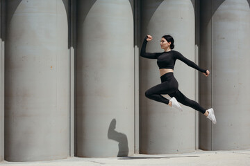 Girl jumping on a background of gray city columns. Street training in a black tracksuit and white sneakers.