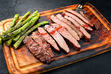 Traditional barbecue sliced dry aged wagyu flank steak offered with green asparagus and chili powder as close-up on a rustic burnt wooden cutting board