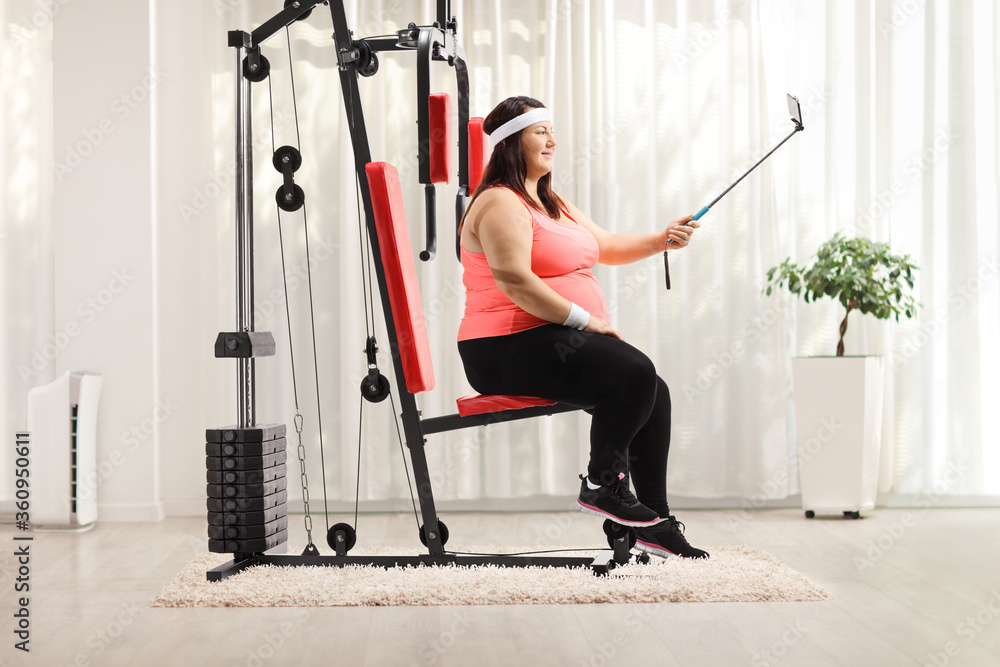 Wall mural overweight woman sitting on a fitness machine and taking a selfie with a stick